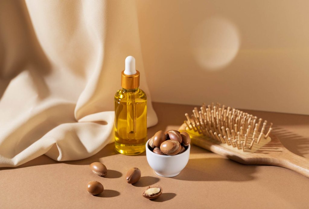Huile cosmétique naturelle à base d’argan, bol de noix, brosse en bois et tissu beige — produits naturels Niya Tijara par Dr Make-up.