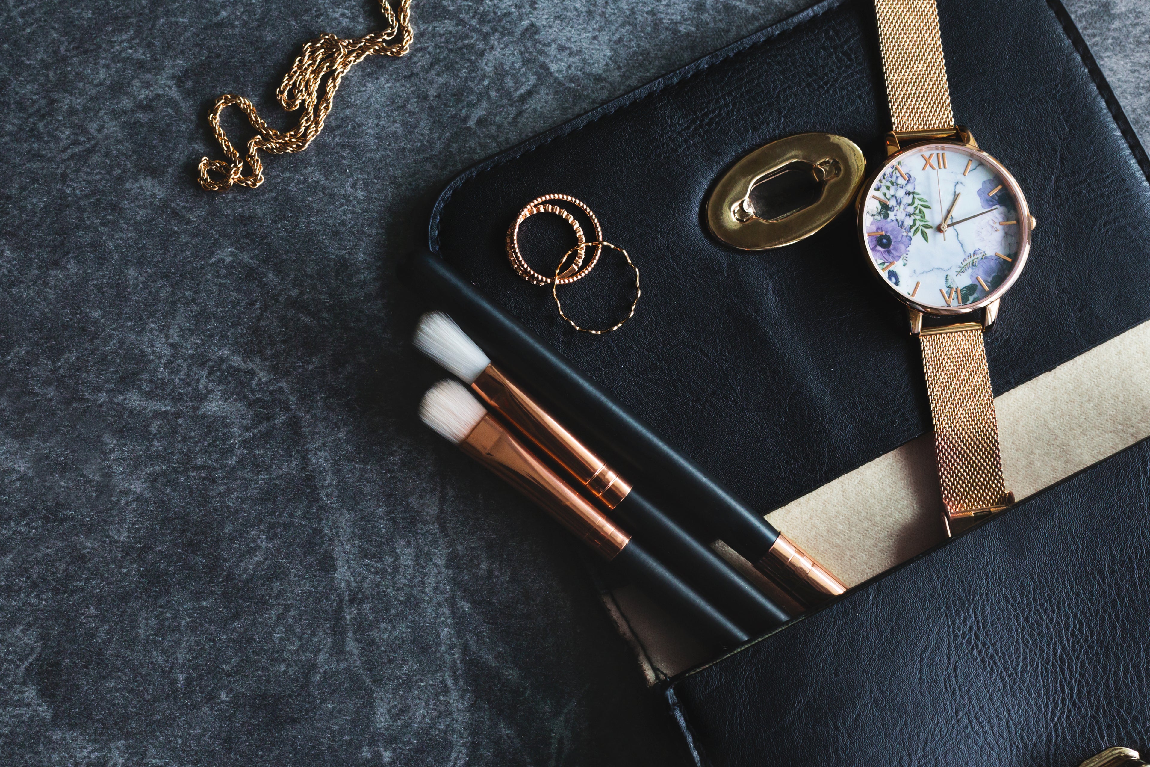 Trousse noire avec pinceaux de maquillage noirs et dorés, montre florale et bijoux dorés sur fond en pierre sombre — visuel élégant de Dr Make-up.